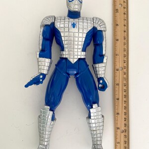 K&ouml;nnte beinhalten: Blaue und silberne Actionfigur von Spider-Man, der einen metallischen Anzug tr&auml;gt. Die Figur ist etwa 15 cm gro&szlig; und steht neben einem Lineal.