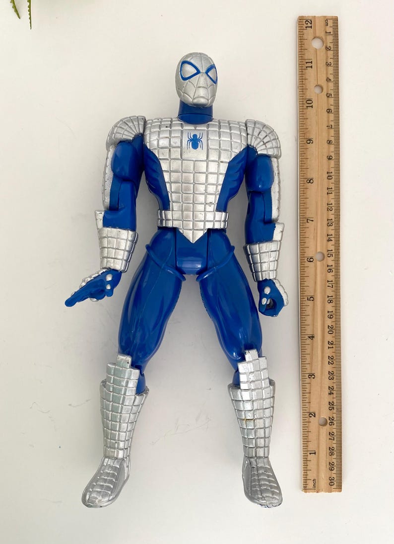 K&ouml;nnte beinhalten: Blaue und silberne Actionfigur von Spider-Man, der einen metallischen Anzug tr&auml;gt. Die Figur ist etwa 15 cm gro&szlig; und steht neben einem Lineal.