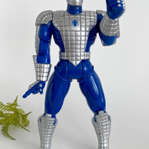 K&ouml;nnte beinhalten: Eine blaue und silberne Actionfigur von Spider-Man, die eine silberne R&uuml;stung tr&auml;gt. Die Figur hat ein Spinnenlogo auf der Brust und ist mit erhobenem rechten Arm in einer Daumen-hoch-Geste posiert.