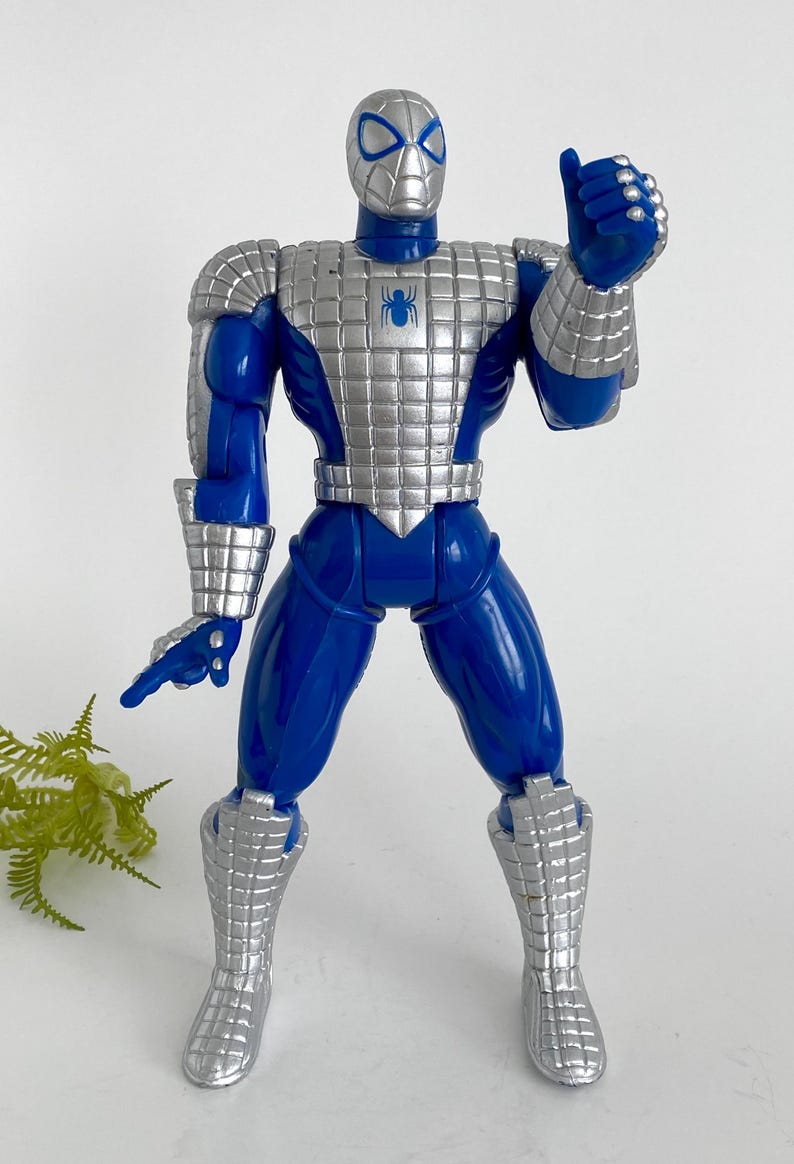 K&ouml;nnte beinhalten: Eine blaue und silberne Actionfigur von Spider-Man, die eine silberne R&uuml;stung tr&auml;gt. Die Figur hat ein Spinnenlogo auf der Brust und ist mit erhobenem rechten Arm in einer Daumen-hoch-Geste posiert.
