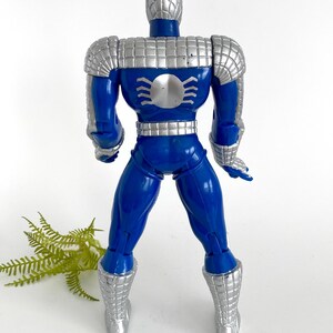 K&ouml;nnte beinhalten: Eine blaue und silberne Actionfigur von Spider-Man, die eine silberne R&uuml;stung tr&auml;gt. Die Figur ist mit den Armen an den Seiten und leicht gespreizten Beinen aufgestellt.