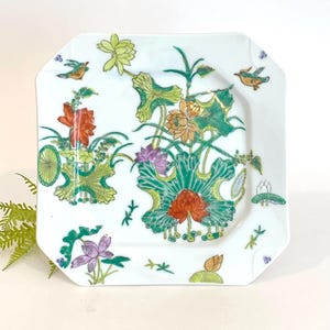 May include: A white square ceramic plate with a green, yellow, orange, and purple floral design. The plate has a slightly octagonal shape and features two small birds on the top corners.