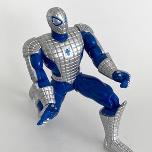 K&ouml;nnte beinhalten: Eine blaue und silberne Actionfigur von Spider-Man in einer knienden Pose. Die Figur tr&auml;gt einen silbernen Anzug mit einem blauen Spinnenlogo auf der Brust.