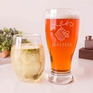 May include: Two clear glasses with a beehive and bee design etched on the front. The text "[BUZZED]" is below the design. One glass is filled with a light yellow liquid and the other is filled with a dark amber liquid.