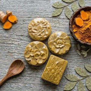 May include: Three round and one rectangular bar of mustard-colored soap with embossed floral patterns. The set includes raw turmeric root pieces, a wooden spoon, and a dark wooden bowl filled with turmeric powder, all arranged on a gray surface.