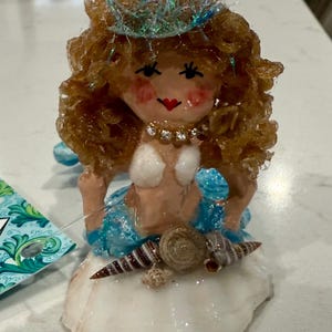 May include: A handcrafted mermaid figurine with brown hair, a blue crown, and a seashell base. The mermaid wears a white top and a blue skirt. Decorative seashells are attached to the base. A unique, whimsical art piece.