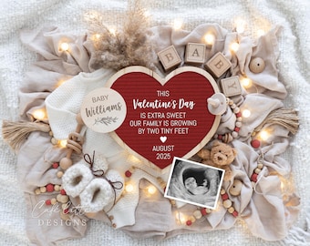 以下が含まれることがあります： 「This Valentine's Day is extra sweet our family is growing by two tiny feet August 2025」というテキストが書かれた赤いハート型のレターボード。ボードは白い毛布、木製のブロック、ベビーラトル、ベビーロンパース、小さなテディベアで囲まれています。