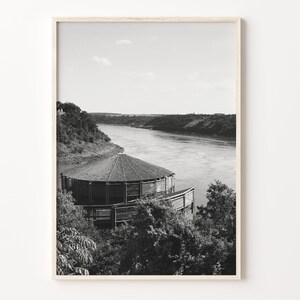 May include: Black and white photograph of a wooden structure with a circular roof overlooking a river. The structure is surrounded by trees and the river is in the background.