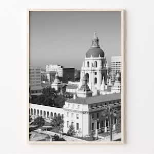 May include: Black and white photograph of the Los Angeles City Hall building. The building has a large dome and is surrounded by other buildings. The photo was taken from a high vantage point.