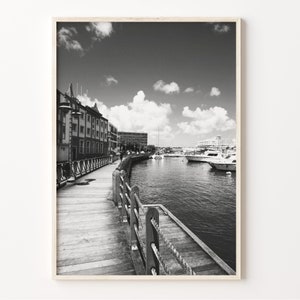 May include: A black and white photograph of a wooden boardwalk leading to a harbor with boats docked. The boardwalk is lined with a wooden railing and there are buildings in the background.