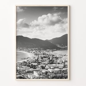 May include: Black and white photograph of a coastal town with a mountain range in the background. The town is built on a peninsula with a beach and ocean in the foreground.