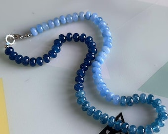 Puede incluir: Un collar compuesto por cuentas azules graduadas, que van desde un azul marino intenso hasta un azul cielo más claro. Las cuentas están ensartadas con un cierre plateado. El collar se exhibe sobre una superficie blanca con formas geométricas.
