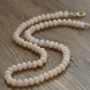 Op de afbeelding: Een witte kralenketting met een gouden sluiting. De ketting is gemaakt van ronde, gladde kralen en ligt op een houten oppervlak.