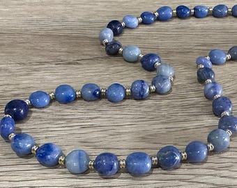 May include: A beaded necklace featuring various shades of blue, from deep navy to light sky blue. The beads are round and interspersed with small silver-colored spacers. The necklace is displayed on a wooden surface.