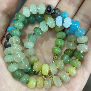 May include: A necklace composed of various shades of green, blue, and yellow beads. The beads are strung together with gold-colored accents and a gold clasp. The beads are oval-shaped and vary in size.