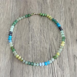 May include: A beaded necklace with a variety of colorful gemstones. The necklace features a gradient of colors, including green, blue, and yellow. The beads are strung together with a gold clasp. The necklace is displayed on a wooden surface.