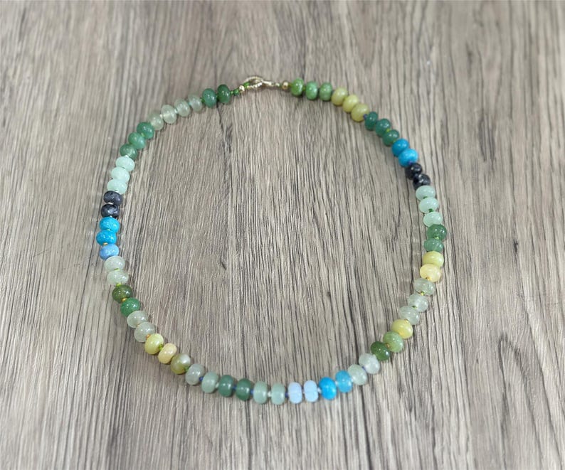 May include: A beaded necklace with a variety of colorful gemstones. The necklace features a gradient of colors, including green, blue, and yellow. The beads are strung together with a gold clasp. The necklace is displayed on a wooden surface.