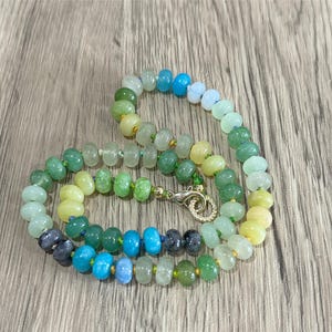 May include: A beaded necklace with a variety of colorful beads. The beads are in shades of green, blue, yellow, and clear. The necklace has a gold-colored clasp. The beads are approximately 0.5 cm in diameter.