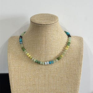 May include: A beaded necklace with a variety of colors, including green, yellow, blue, and black. The necklace is displayed on a tan mannequin. The beads are small and round, and the necklace appears to be handmade.