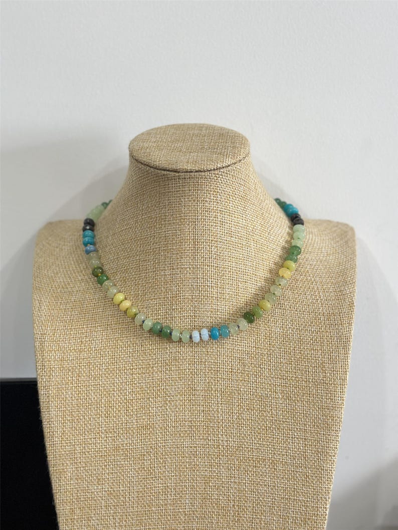 May include: A beaded necklace with a variety of colors, including green, yellow, blue, and black. The necklace is displayed on a tan mannequin. The beads are small and round, and the necklace appears to be handmade.