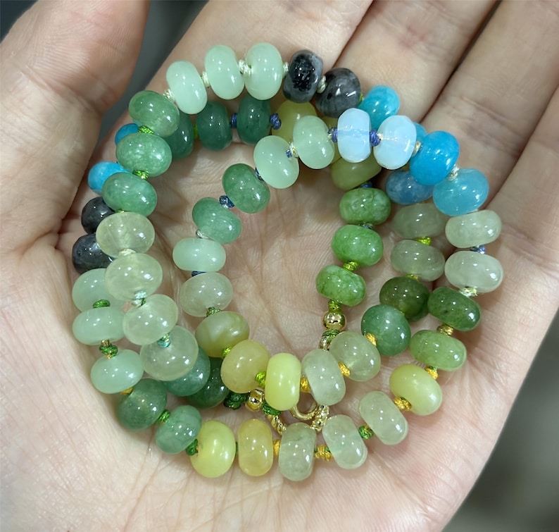May include: A necklace composed of various shades of green, blue, and yellow beads. The beads are strung together with gold-colored accents and a gold clasp. The beads are oval-shaped and vary in size.