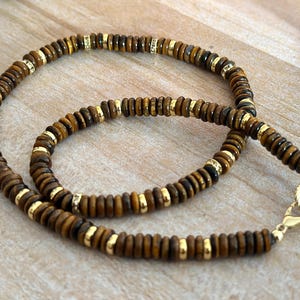 May include: A beaded necklace featuring brown and gold beads. The necklace has a gold clasp and gold accents between the brown beads. The beads are small and round, creating a layered effect. The necklace is displayed on a light-colored wooden surface.