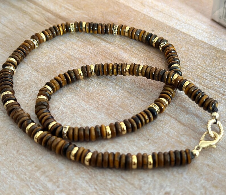 May include: A beaded necklace featuring brown and gold beads. The necklace has a gold clasp and gold accents between the brown beads. The beads are small and round, creating a layered effect. The necklace is displayed on a light-colored wooden surface.