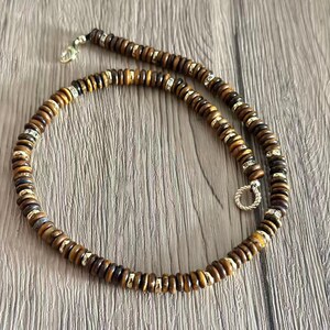 May include: A beaded necklace featuring brown and gold-toned wooden beads. The necklace has silver-toned accents and a clasp. The beads are round and uniform in size, creating a simple yet elegant design. The necklace is displayed on a wooden surface.