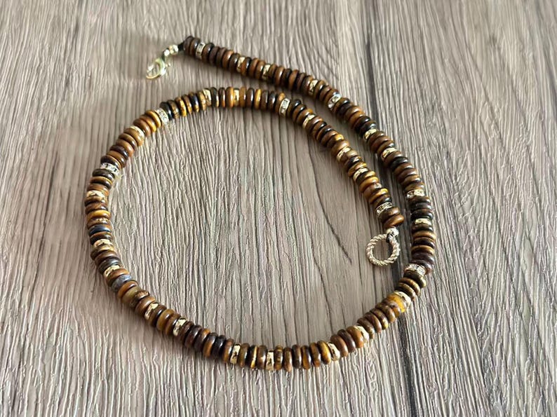 May include: A beaded necklace featuring brown and gold-toned wooden beads. The necklace has silver-toned accents and a clasp. The beads are round and uniform in size, creating a simple yet elegant design. The necklace is displayed on a wooden surface.