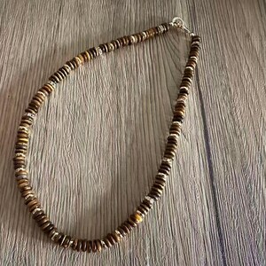 May include: A necklace composed of small, round, brown beads with silver-colored spacers. The beads are strung together to form a necklace, and the clasp is silver-colored. The necklace is displayed on a wooden surface.
