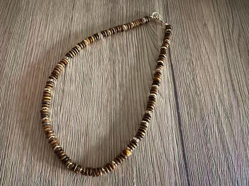 May include: A necklace composed of small, round, brown beads with silver-colored spacers. The beads are strung together to form a necklace, and the clasp is silver-colored. The necklace is displayed on a wooden surface.