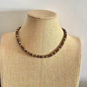 May include: A brown beaded necklace with alternating dark and light brown beads, displayed on a tan mannequin. The necklace has a simple, circular design, suitable for casual wear.