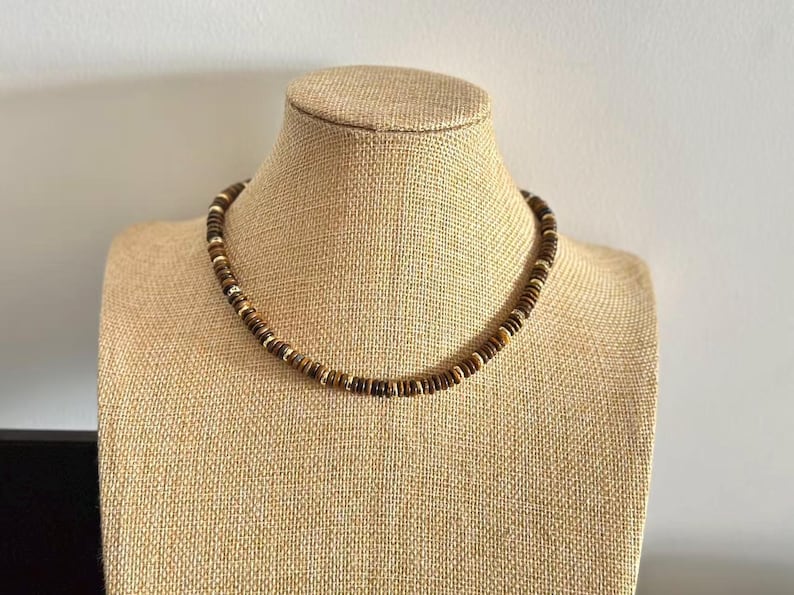 May include: A brown beaded necklace with alternating dark and light brown beads, displayed on a tan mannequin. The necklace has a simple, circular design, suitable for casual wear.