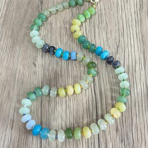 May include: A colorful beaded necklace with a gradient of colors, including green, blue, and yellow. The necklace is made of small, round beads and has a gold clasp. The beads are strung together on a thin, clear cord.