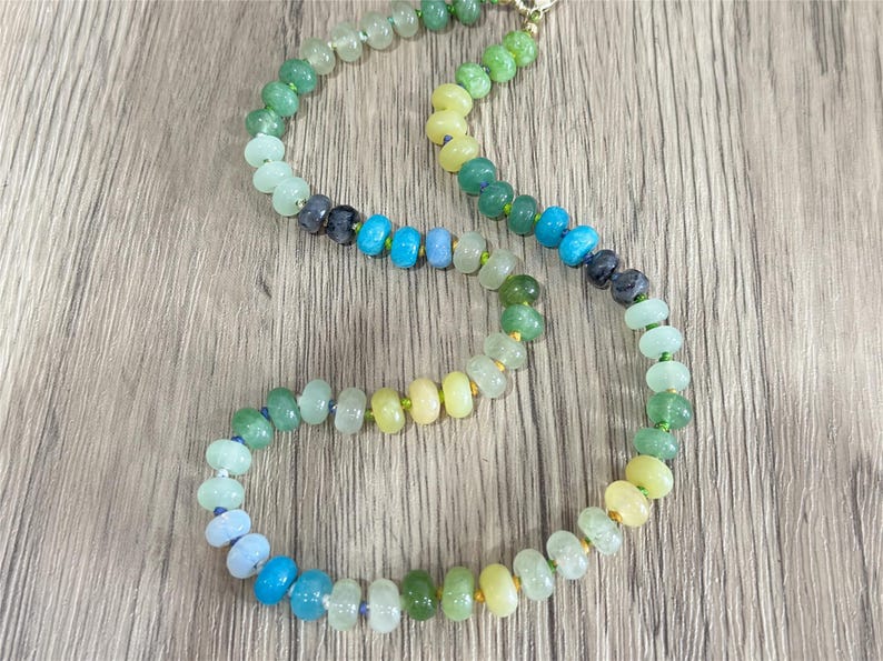 May include: A colorful beaded necklace with a gradient of colors, including green, blue, and yellow. The necklace is made of small, round beads and has a gold clasp. The beads are strung together on a thin, clear cord.