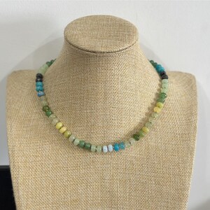 May include: A beaded necklace with a variety of colors, including green, yellow, blue, and black. The necklace is displayed on a tan jewelry bust. The beads are small and round, and the necklace appears to be handmade.