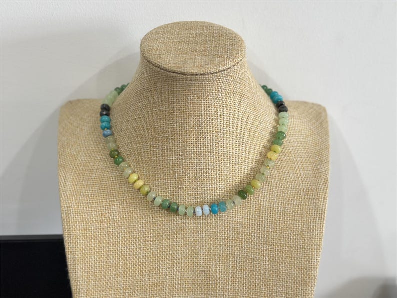 May include: A beaded necklace with a variety of colors, including green, yellow, blue, and black. The necklace is displayed on a tan jewelry bust. The beads are small and round, and the necklace appears to be handmade.