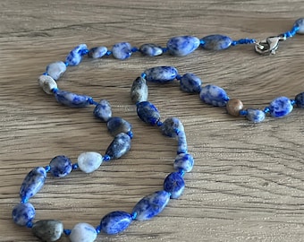 May include: A necklace featuring blue and white oval-shaped beads strung on a blue cord. The beads have a marbled appearance and are connected with a silver clasp. The necklace is displayed on a wooden surface.