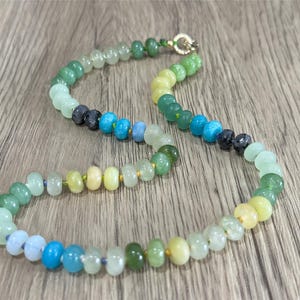 May include: A colorful beaded necklace featuring a variety of gemstones in shades of green, blue, and yellow. The necklace has a gold clasp and is displayed on a wooden surface. The beads are round and vary in size.