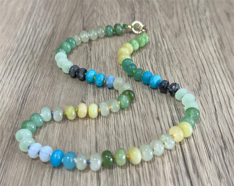 May include: A colorful beaded necklace featuring a variety of gemstones in shades of green, blue, and yellow. The necklace has a gold clasp and is displayed on a wooden surface. The beads are round and vary in size.