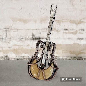 May include: A decorative guitar sculpture crafted from coiled copper wire and a smooth, tan-colored stone body. The instrument features a long, slender neck with wire-wrapped frets and a headstock, all constructed from metallic copper wire.