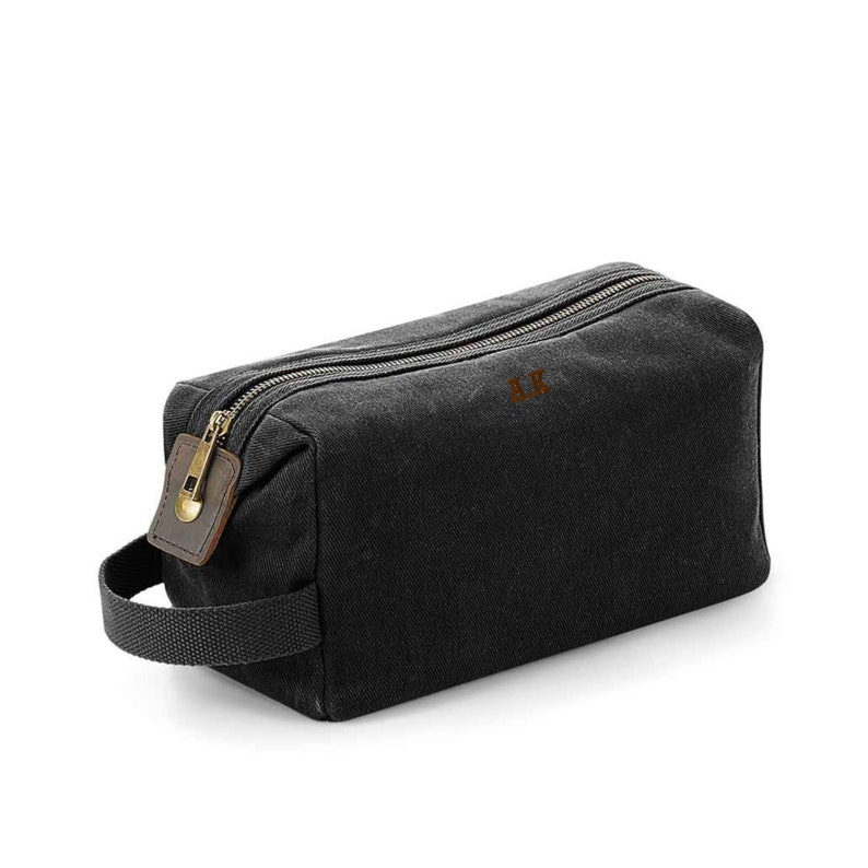 May include: A black canvas wash bag with a brown leather zip pull and a gold zip. The bag has a black strap handle and the initials "A.K." embroidered on the front.