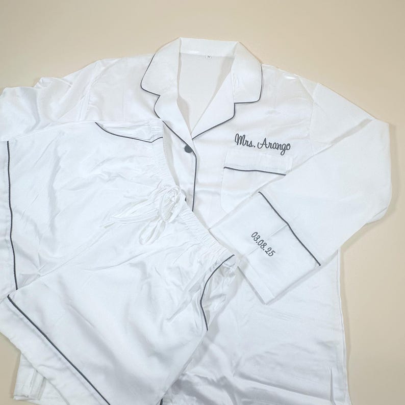 May include: White pajama set with black trim, including a button-down shirt and shorts. The shirt is embroidered with "Mrs. Arango" and "03.08.25" on the cuff. The set is made of a smooth, silky material.