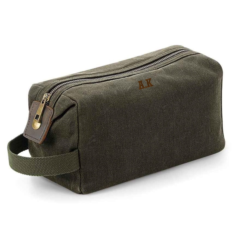 May include: A green canvas wash bag with a brown leather zip pull and a brown leather handle. The bag has a zip closure and the initials "AK" are embroidered on the front.