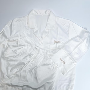 May include: White satin pajama set with a collared button-down shirt and drawstring pants. The shirt is embroidered with the name "Jessica" and the date "26.05.2024" on the cuff. The set is made of a shiny, smooth fabric.