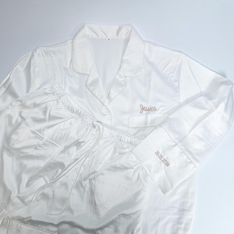 May include: White satin pajama set with a collared button-down shirt and drawstring pants. The shirt is embroidered with the name "Jessica" and the date "26.05.2024" on the cuff. The set is made of a shiny, smooth fabric.