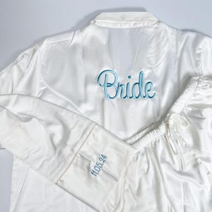 May include: White satin pajama set with light blue embroidered text that reads "Bride" on the back of the top. The sleeve cuff has the date "11.05.24" embroidered in light blue. The set includes a button-down top and drawstring pants.