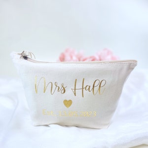 May include: A white canvas makeup bag with a gold zipper and gold lettering that says "Mrs. Hall Est. 13.05.2023" with a gold heart symbol.