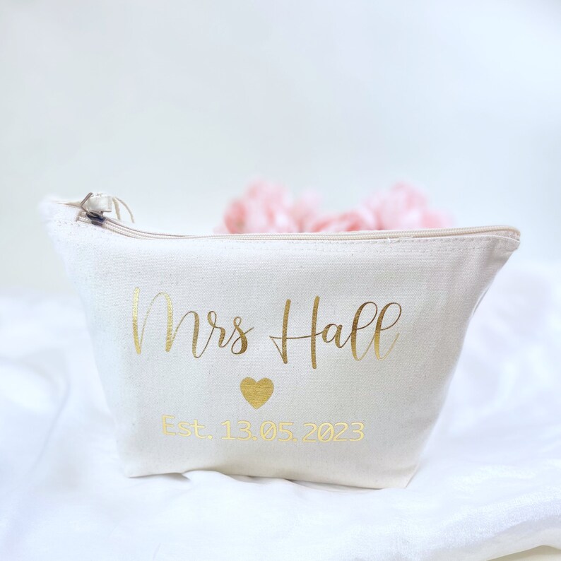 May include: A white canvas makeup bag with a gold zipper and gold lettering that says "Mrs. Hall Est. 13.05.2023" with a gold heart symbol.