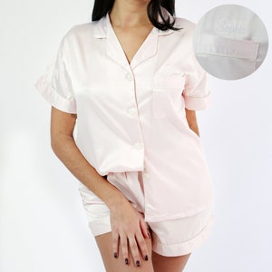 May include: A pink satin pyjama set with short sleeves and shorts. The top has a button-up front and a pocket on the left side. The shorts have a button and zip closure. The top has the name "Sophie" embroidered on the pocket.